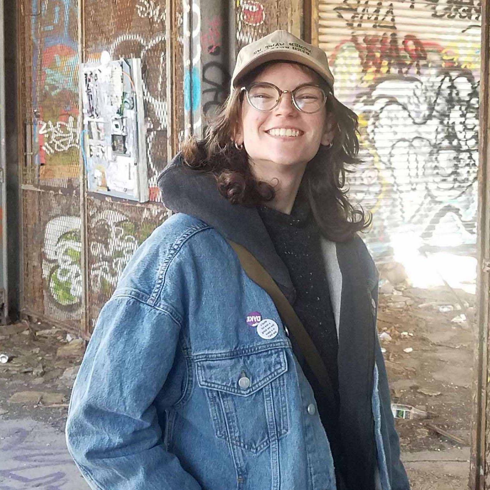 Tori smiles in a jean jacket before a graffiti wall